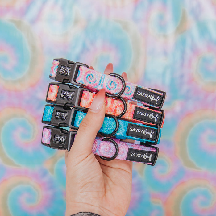 A hand displays five Sassy Woof “Dog Collar - Tropical Tides” pieces with pink, blue, and purple tie dye on neoprene. The adjustable collars appear against a pastel rainbow swirl backdrop for a playful look.