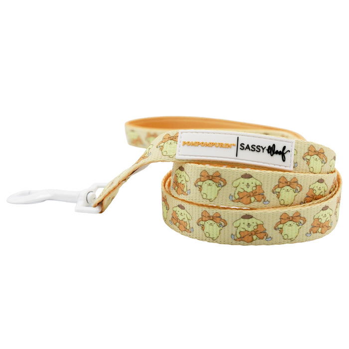 Charming Sassy Woof Dog Collar Three Piece Bundle - Pompompurin™ featuring a yellow design with repeating cartoon Pompompurin characters, tan trim, a white clip, and the Sassy Woof brand label. This adorable Sanrio dog accessory set is coiled with the clasp visible in the foreground.