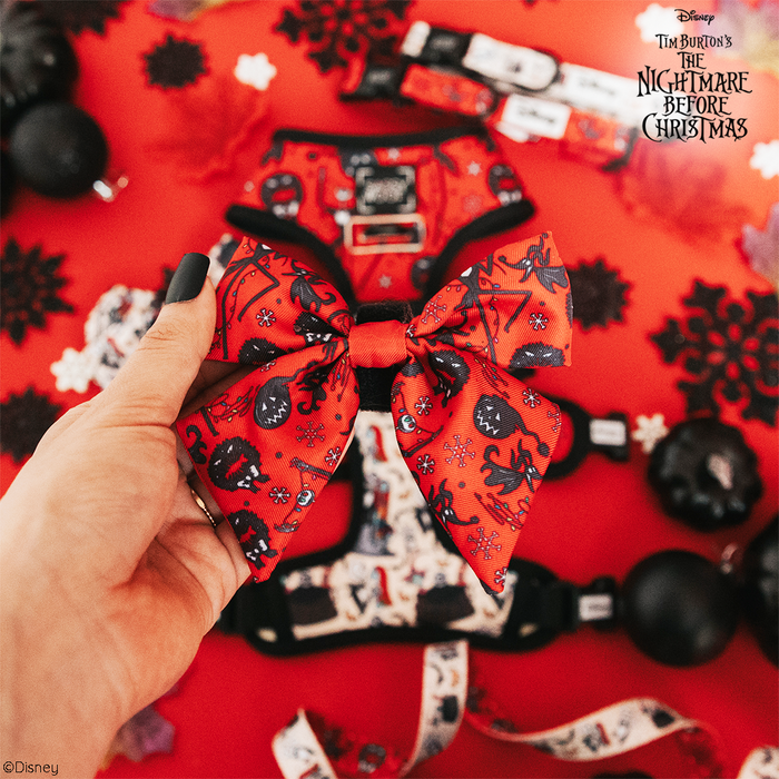 A hand displays the Sassy Woof Dog Sailor Bow - The Nightmare Before Christmas™ (Red), featuring themed patterns. Matching adjustable harness pet accessories and decorations are arranged on a red surface, with the movie title in the top right corner.