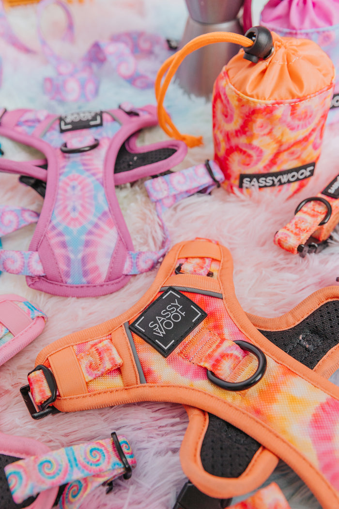 Sassy Woof’s Dog Step-in Harness in Summer Sorbet, featuring orange and pink tie-dye with reflective straps, is displayed on a pink furry surface among other colorful pastel pet accessories.