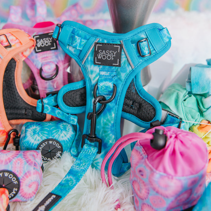 A Sassy Woof Dog Step-in Harness - Tropical Tides and matching leash are featured among colorful dog accessories, like a pink harness and tie-dye waste bag holders, displayed on a fluffy white surface.