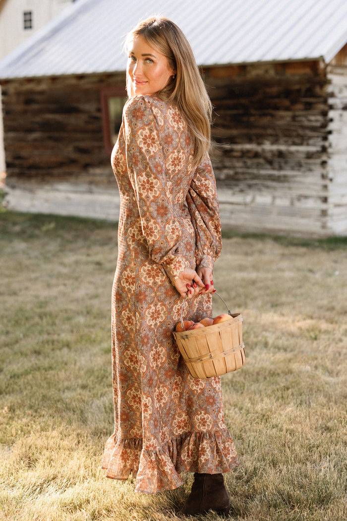 A woman wearing the Ivy City Co Draper Dress in Golden Hour Tesserae, featuring elegant balloon sleeves and a long, floral design, stands on the grass holding a basket of apples and looking back over her shoulder. An old wooden building can be seen in the background.