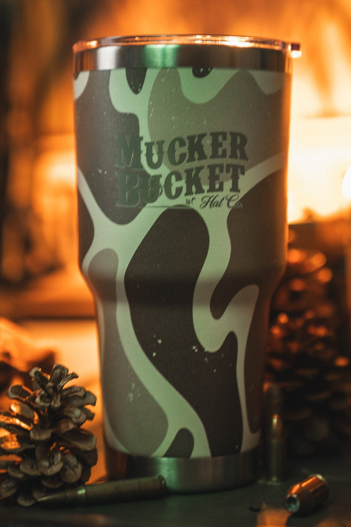 The Duck Camo Tumbler - 30oz by Mucker Bucket features a green and tan camouflage design and custom laser engraving, displayed with pinecones, a bullet, and warm ambient lighting.