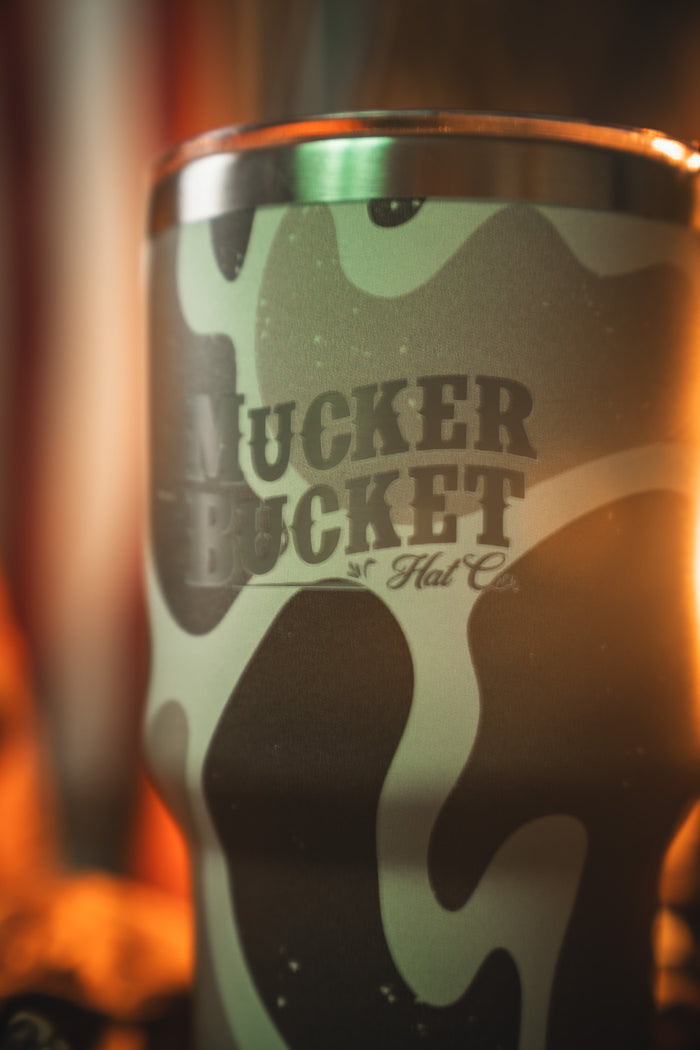 A close-up of the Mucker Bucket Duck Camo Tumbler - 30oz, with "MUCKER BUCKET" boldly on its laser-engraved stainless steel. Warm light enhances the unique camouflage pattern.