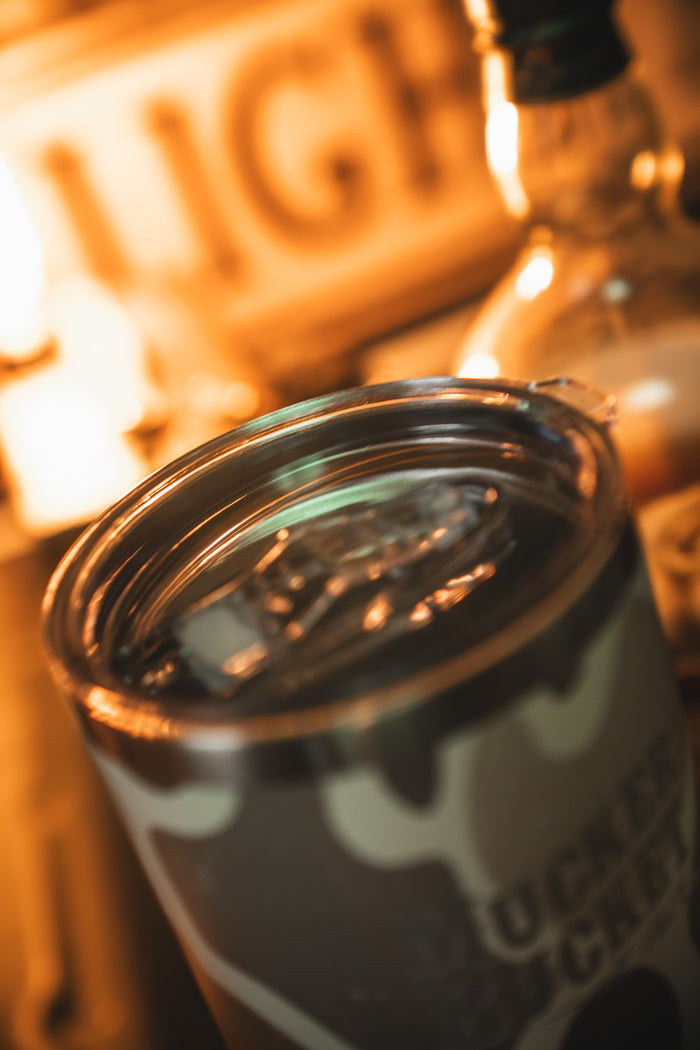 A close-up of the Mucker Bucket Duck Camo Tumbler - 30oz filled with ice and a drink, with a glowing LIGHT sign and bottle blurred in the background—a custom laser-engraved stainless steel tumbler for any setting.