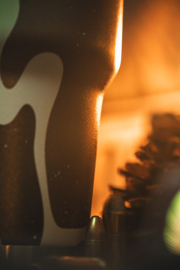 Close-up of the Mucker Bucket Duck Camo Tumbler - 30oz, illuminated by warm orange light, with a pinecone and bullet nearby. Custom laser engraving makes this rugged stainless steel tumbler uniquely yours.