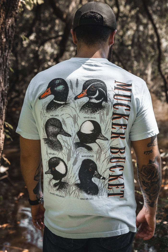 A man in a cap stands by a wooded stream, wearing the Duck DNA Premium Tee by Mucker Bucket—a relaxed fit light blue shirt featuring illustrations and names of five duck species with “Mucker Bucket” printed vertically on the back.