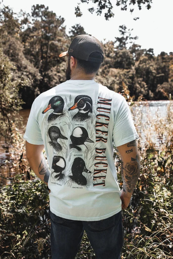 A man stands by a river wearing the Duck DNA Premium Tee from Mucker Bucket, featuring duck illustrations and “MUCKER BUCKET” on the back, paired with his tattoos, baseball cap, and a scenic backdrop of trees and water.