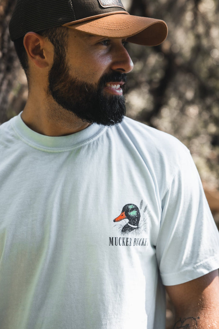 A bearded man in a brown cap smiles outdoors, wearing the Mucker Bucket Duck DNA Premium Tee—a light garment-dyed cotton shirt featuring a duck graphic and the words "Mucker Bucket.