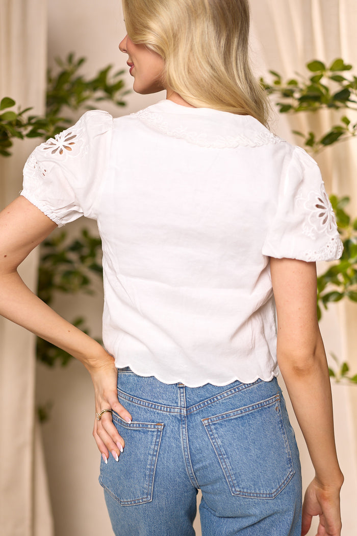 A woman with long blonde hair is seen from the back wearing the Endora Top in Ivory by Ivy City Co—an ivory cotton eyelet top with scalloped edges and floral embroidery—paired with high-waisted blue jeans. Blurred green plants are in the background.