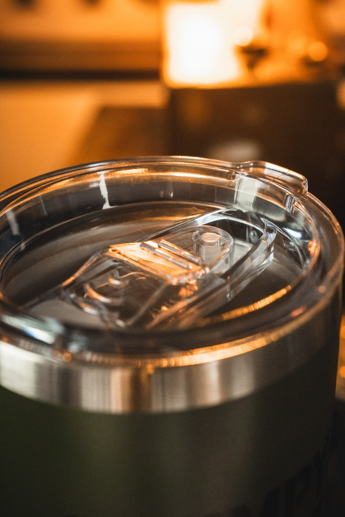 The Grumpy Old Vet Tumbler - 20oz by Born United Private Label features a sleek stainless steel design with a clear lid, filled with a beverage and set against a warmly lit, softly blurred background.