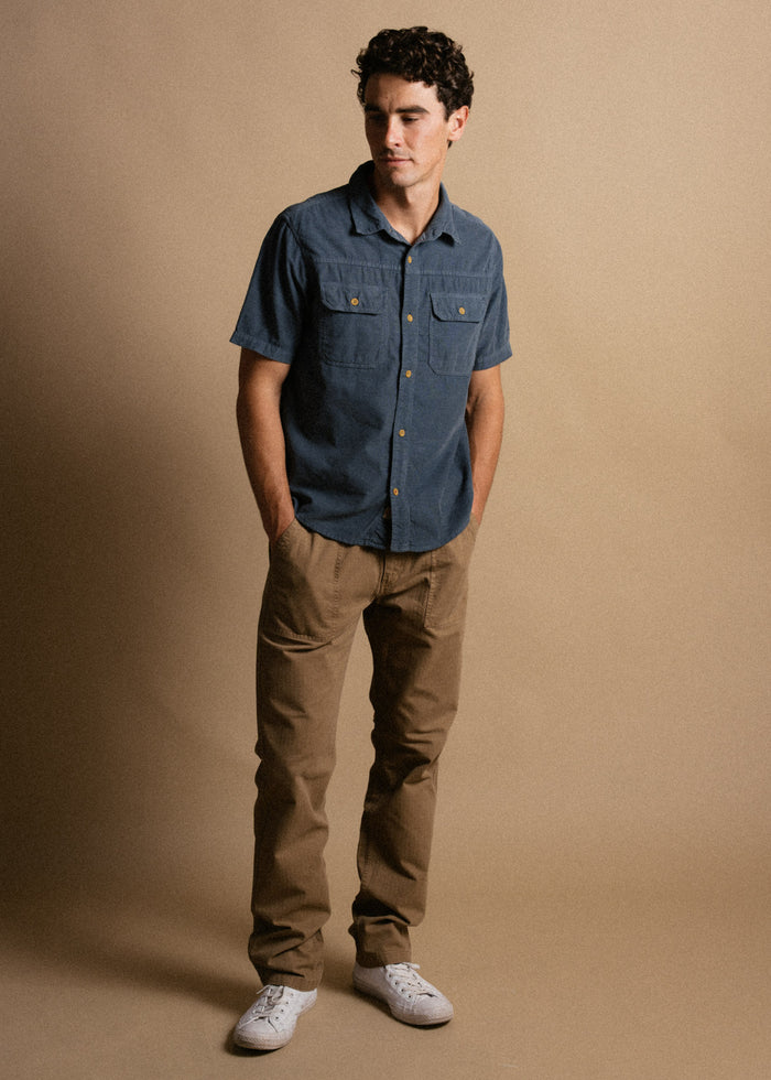 Man in a blue shirt and khaki pants stands against a beige background, hands in pockets, looking down, wearing white sneakers.