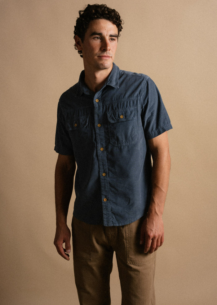 Man in a blue button-up shirt and khaki pants stands against a beige background, looking to the side with a thoughtful expression.