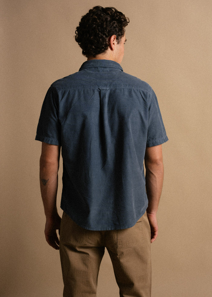 Man in blue shirt and khaki pants stands with back to camera against beige background, showcasing casual style and curly hair.