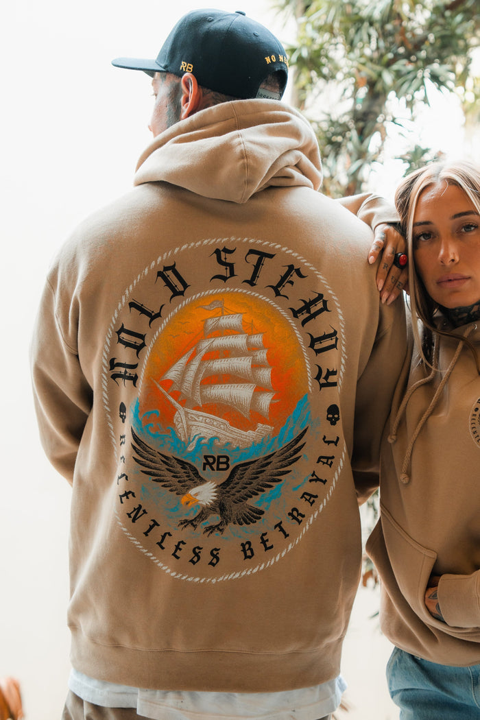 Two people in tan Hold Steady Premium Hoodies by Relentless Betrayal stand side by side, both wearing the unisex heavyweight sweatshirt featuring a bold sailing ship, eagle graphic, and Hold Steady Relentless Betrayal text.