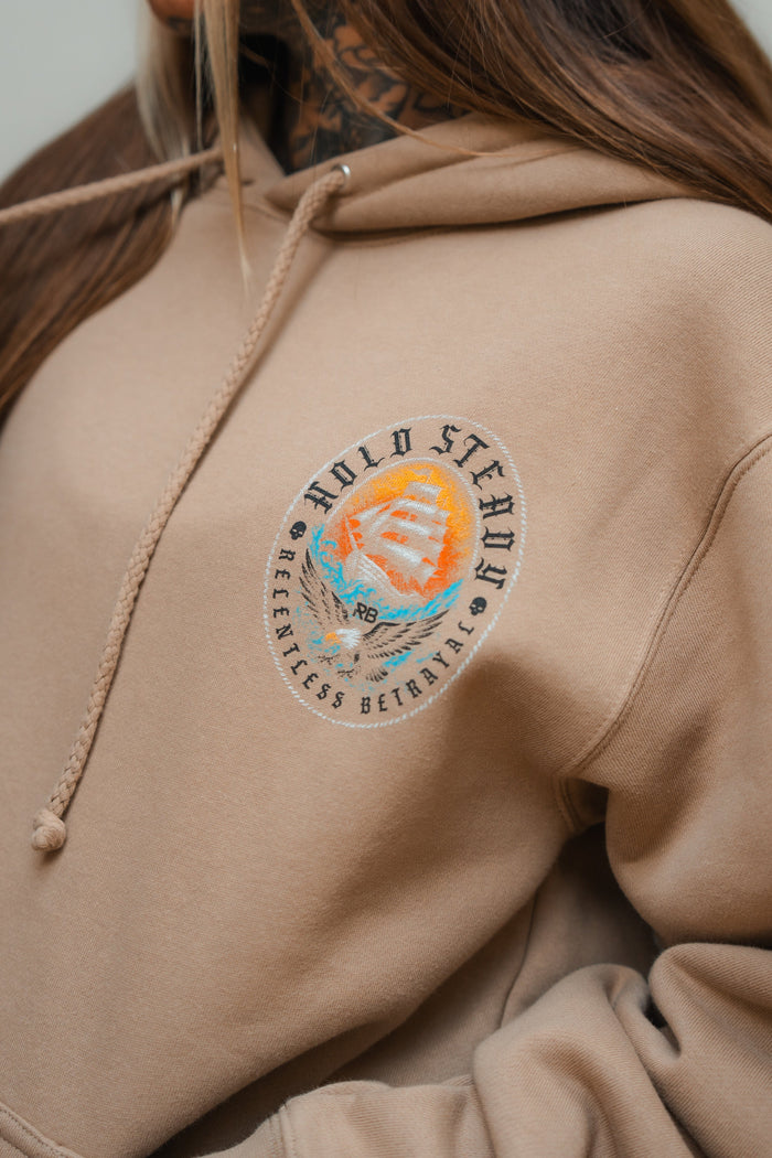 A person wears the Hold Steady Premium Hoodie by Relentless Betrayal, featuring a tan heavyweight fabric and an embroidered sailing ship patch on the chest. Only part of their face and hair is visible, highlighting this unique unisex hoodie.