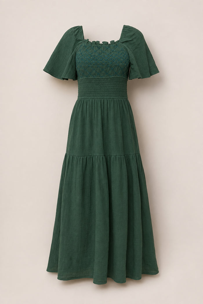 The Jade Dress in Evergreen by Ivy City Co is a dark green, ankle-length cotton dress featuring short flutter sleeves, a smocked square neckline, a fitted waist, and a tiered skirt. Maternity friendly and shown against a plain light beige background.