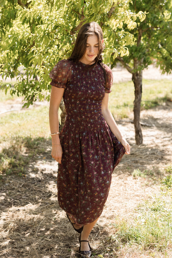 A woman in the Ivy City Co Jennifer Dress in Aubergine Elysian Vine, featuring a smocked bodice and dark floral print, walks outdoors on a sunny day, gracefully holding her skirt. Surrounded by lush green trees and gentle sunlight, she creates a serene, natural scene—showcasing an effortlessly elegant style that’s also maternity friendly.