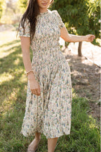 A woman stands outdoors on grass, barefoot and surrounded by trees and sunlight, wearing the Ivy City Co Jennifer Dress in Autumn Bouquet—a light-colored, floral-print, midi-length dress with a smocked bodice and gathered waist. Featuring short sleeves, a relaxed fit, and a maternity-friendly design, this style perfectly complements her long brown hair for an effortlessly graceful look.
