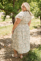 Wearing the Jennifer Dress in Autumn Bouquet by Ivy City Co, a woman stands on a sun-dappled dirt path surrounded by lush green trees, smiling as she looks over her shoulder. With its smocked bodice and floral print, this effortlessly maternity-friendly dress offers comfort and style for enjoying nature on a bright day.