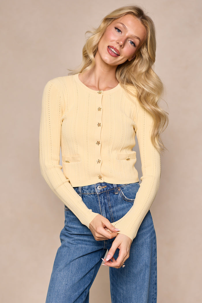 A woman with long blonde hair wears the Ivy City Co Kali Cardigan in Butter Yellow and blue jeans. She stands against a beige background, smiling softly with her hands loosely clasped at her waist.