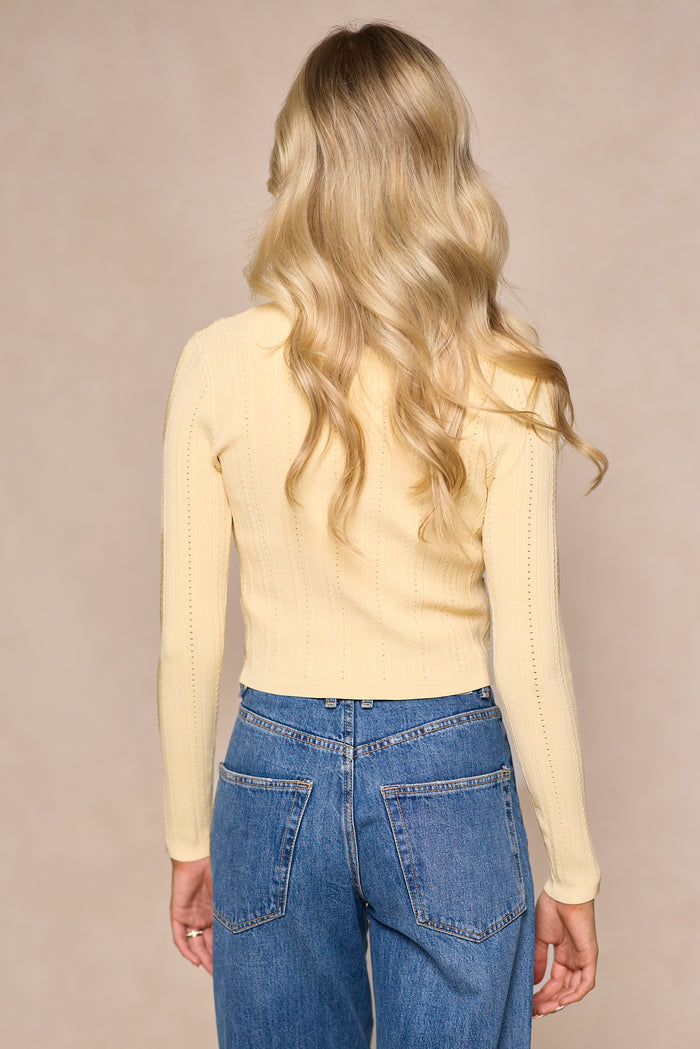 A woman with long, wavy blonde hair is seen from behind wearing the Ivy City Co Kali Cardigan in Butter Yellow as a spring layering piece, paired with high-waisted blue jeans against a neutral beige background.