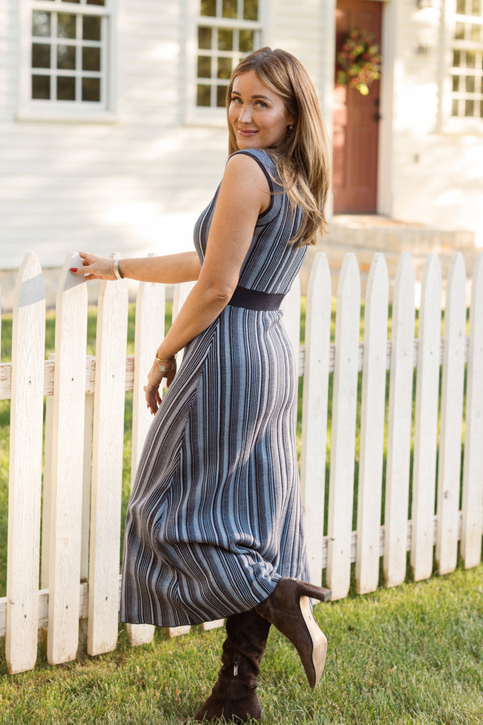 Wearing the Laurel Dress in Midnight by Ivy City Co, a woman stands by a white picket fence in boots, glancing over her shoulder with a smile. The maternity-friendly sweater knit dress flatters her figure as she poses near a white house with windows and a red door in the background.