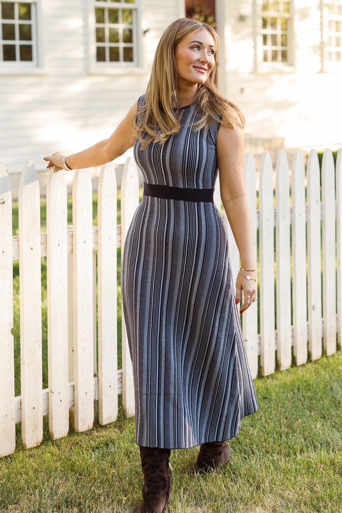 A woman stands on grass by a white picket fence, smiling and looking to the side, wearing the sleeveless, tea-length Laurel Dress in Midnight by Ivy City Co. Sunlight illuminates her while a white house appears in the background.