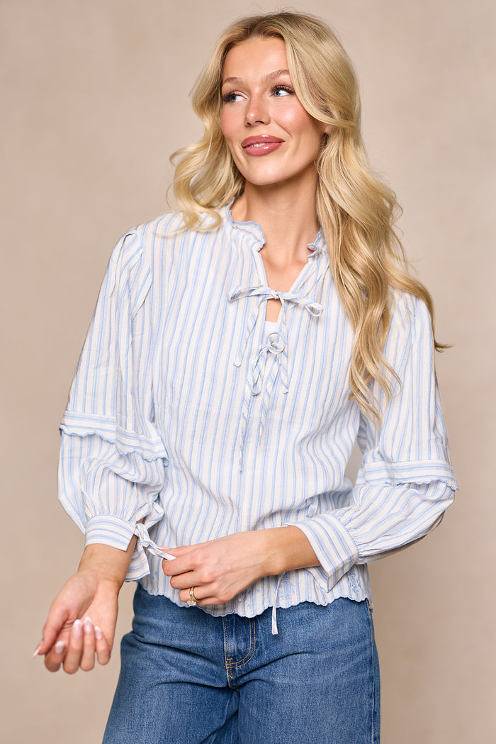 A woman with long blonde hair wears the Ivy City Co Leticia Top in White & Blue Stripe, featuring tie details at the neckline, paired with blue jeans—a stylish choice for your spring wardrobe.