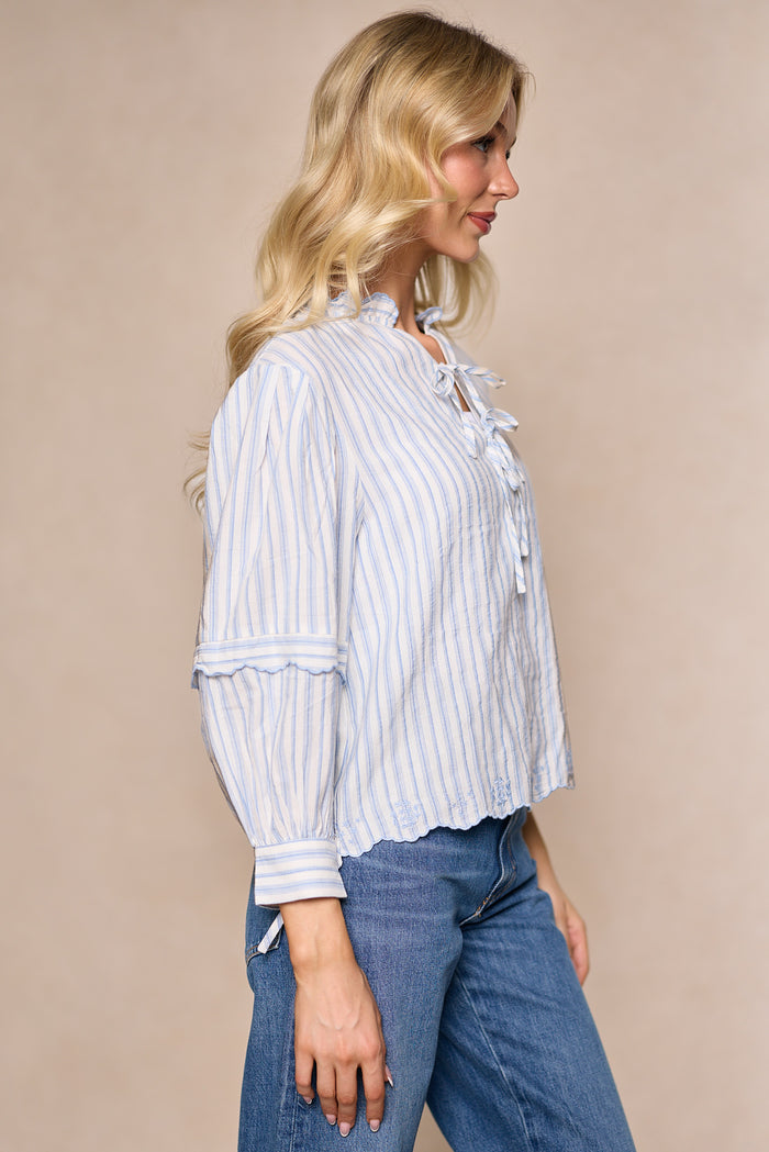 A woman with long blonde hair stands sideways against a beige background, wearing the Ivy City Co Leticia Top in White & Blue Stripe, featuring cotton-poly blend fabric, ruffled details, and blue jeans.