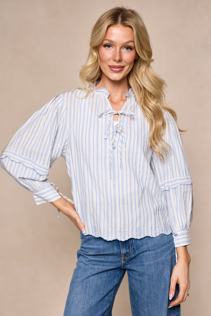A woman with long blonde hair is standing against a beige background and smiling, wearing the Leticia Top in White & Blue Stripe by Ivy City Co—a light blue and white striped spring blouse featuring a tie detail at the neck. Crafted from a comfortable cotton-poly blend, this top creates a flattering silhouette that’s perfect for the season, paired here with blue jeans.