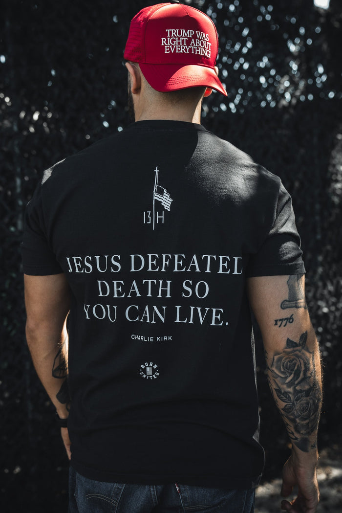 A man with tattooed arms faces away from the camera, wearing a red TRUMP WAS RIGHT ABOUT EVERYTHING cap and a Born United Private Label "Live Like Charlie" t-shirt that reads JESUS DEFEATED DEATH SO YOU CAN LIVE. CHARLIE KIRK.