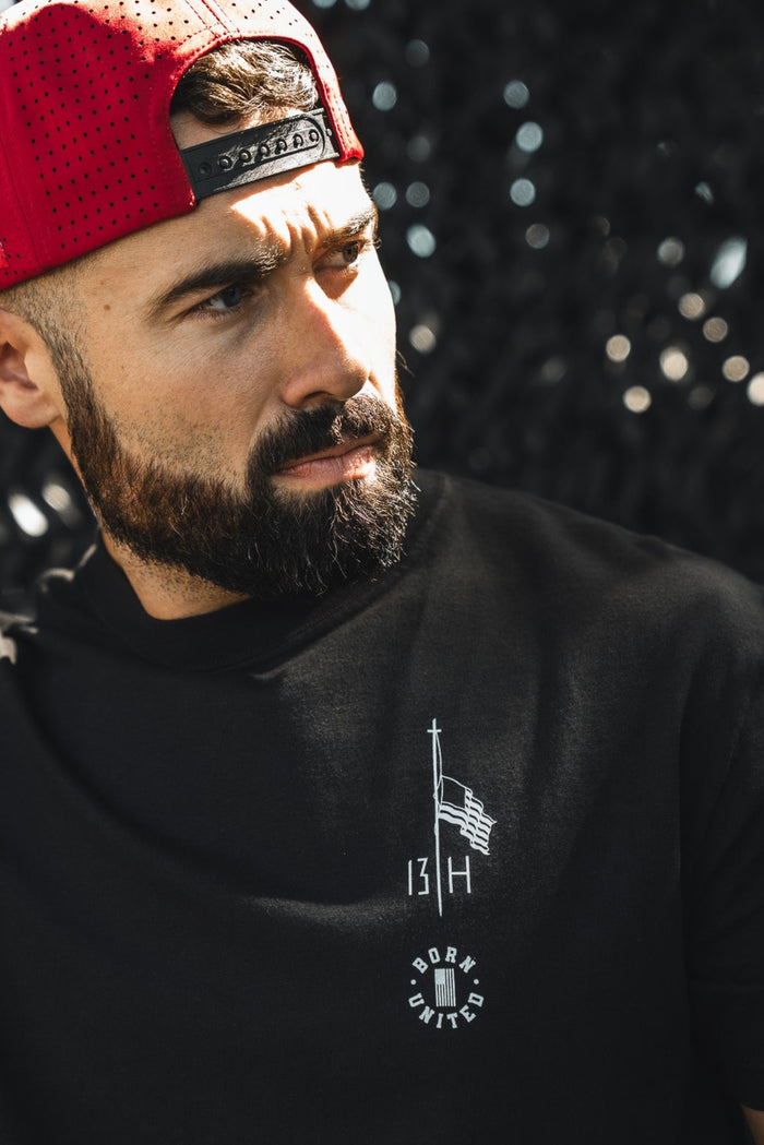 A bearded man in a red backward baseball cap and a Born United Private Label Live Like Charlie tee stands in sunlight, adjusting his hat, with a dark textured background behind him.