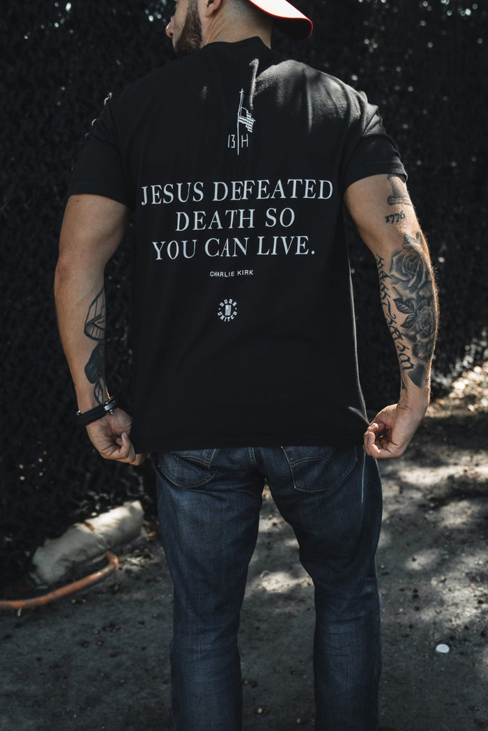A man stands outdoors with his back to the camera, wearing jeans, a red backwards cap, and a Born United Private Label "Live Like Charlie" t-shirt featuring the message "Jesus defeated death so you can live.