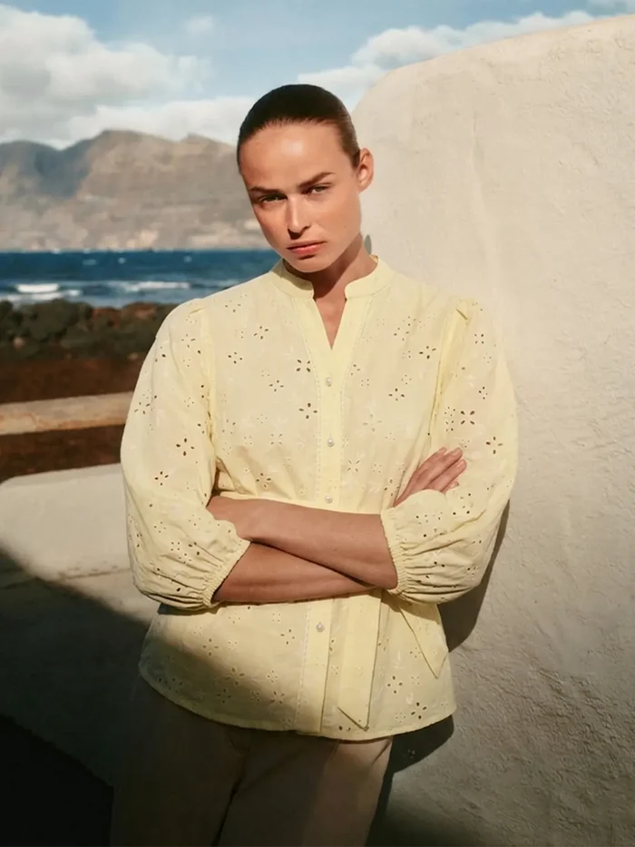 Woman in a yellow blouse standing against a scenic background with mountains and ocean.