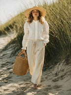 Woman in a white outfit with a straw hat and bag on a sandy beach.