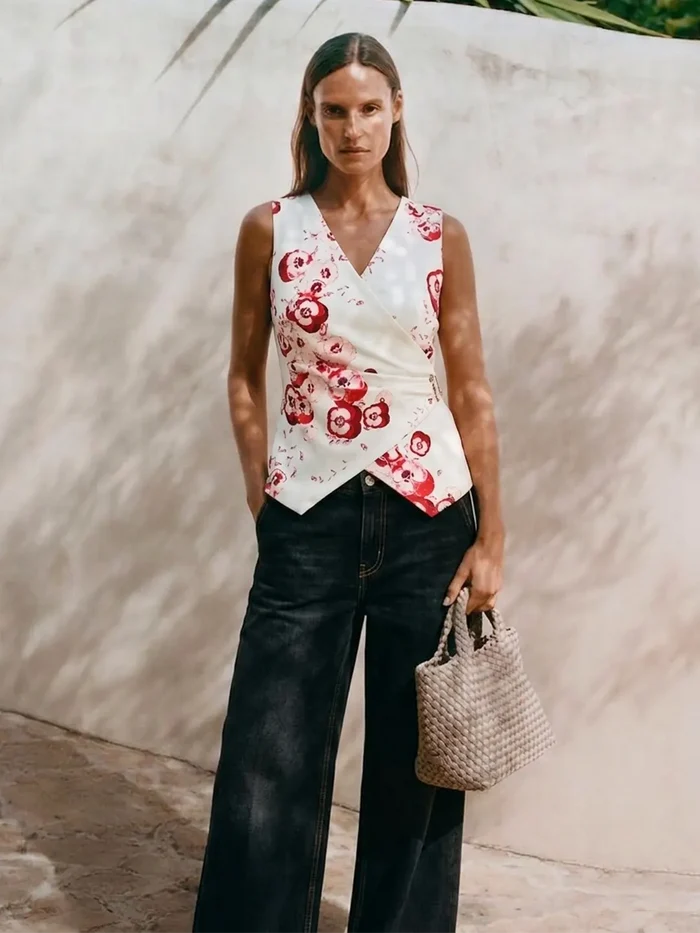 Woman wearing a floral sleeveless top and black pants, holding a woven handbag against a neutral background.