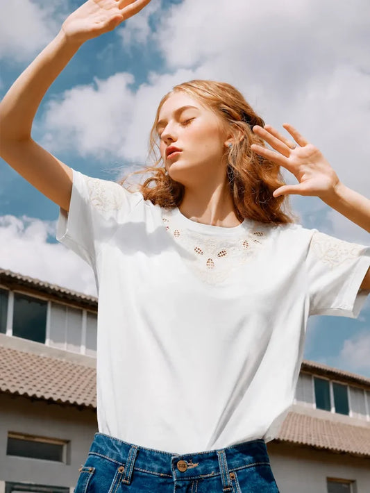 Woman wearing a white t-shirt with lace details and blue jeans, standing outdoors with a building in the background.