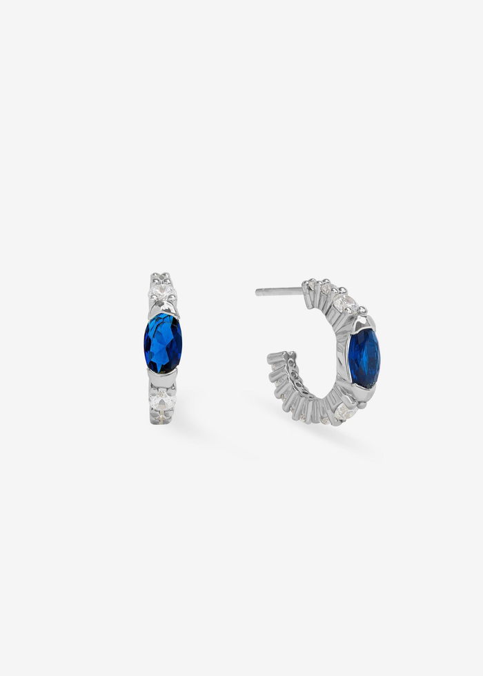 A pair of silver huggies with a blue sapphire stone against a white background