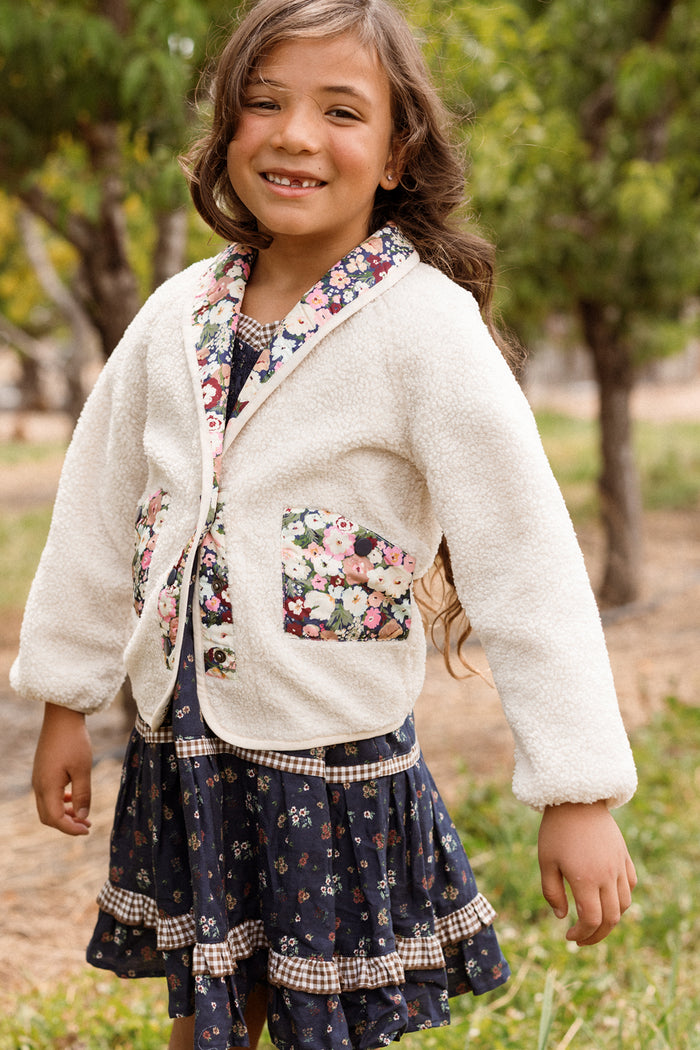 A smiling child with long hair stands outdoors amidst trees and greenery, wearing a floral dress topped with the Ivy City Co Mini Britta Jacket in Ivory & Midnight Garden. This charming jacket features balloon sleeves, floral trim, and pockets.