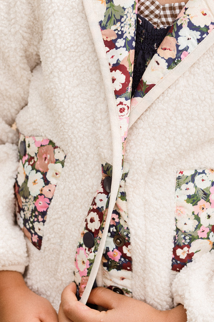 An individual wears the Mini Britta Jacket in Ivory & Midnight Garden by Ivy City Co—a white, fluffy sherpa jacket accented with floral-patterned trim and pockets, plus balloon sleeves—holding its front edges together.