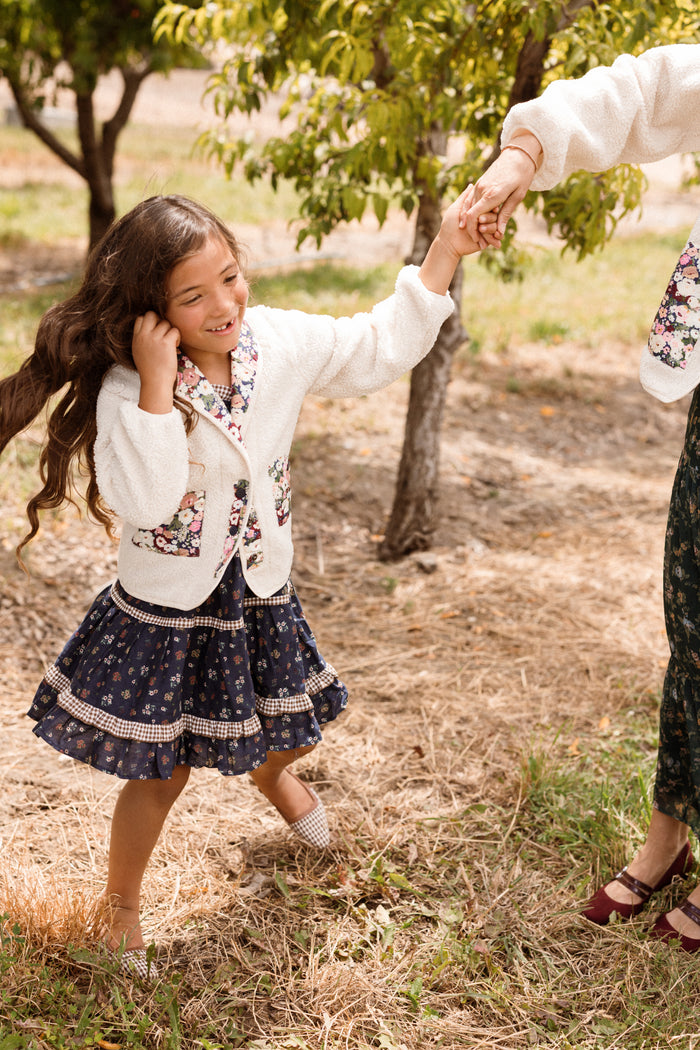 A young girl beams while holding an adult’s hand outdoors near trees on a sunny day, wearing the Ivy City Co Mini Britta Jacket in Ivory & Midnight Garden with its playful balloon sleeves.
