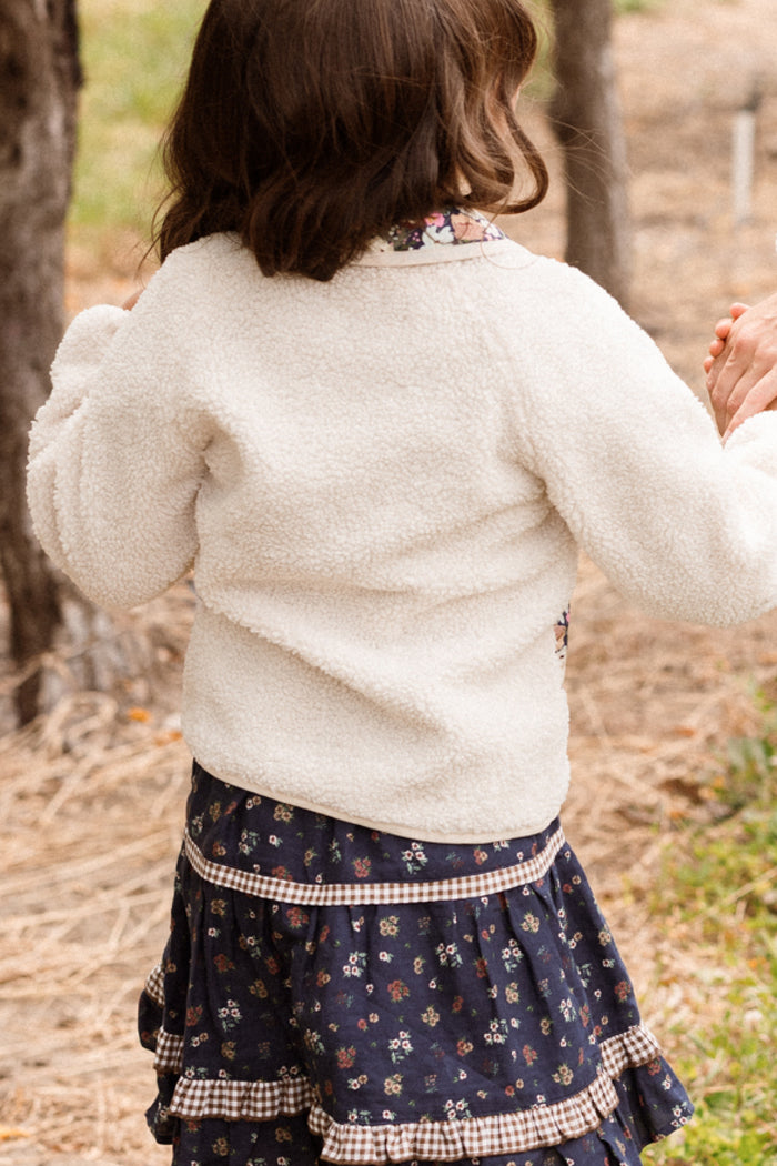 A child with brown hair stands outdoors on dry grass, facing away from the camera, wearing the Ivy City Co Mini Britta Jacket in Ivory paired with a Midnight Garden floral skirt.