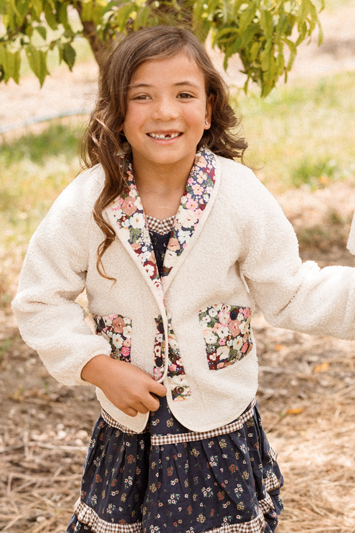 A smiling young girl with long brown hair is wearing the Ivy City Co Mini Britta Jacket in Ivory & Midnight Garden, featuring floral trim and balloon sleeves, over a patterned navy dress as she stands outdoors in a garden or park.