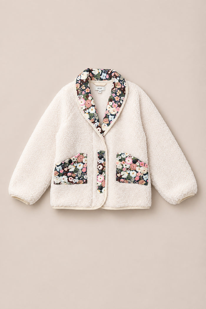 The Mini Britta Jacket in Ivory & Midnight Garden by Ivy City Co features a cozy cream sherpa exterior with a fuzzy texture, accented by floral fabric details on the collar, pocket linings, and front opening. Designed with two front pockets and statement balloon sleeves, this jacket effortlessly combines warmth and style.