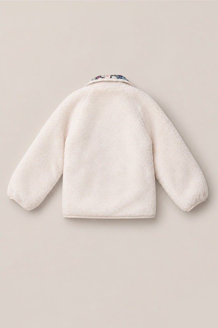The Mini Britta Jacket in Ivory & Midnight Garden by Ivy City Co is displayed laid flat with the back facing up. This cozy sherpa jacket features long balloon sleeves and a collar accented with a subtle midnight garden floral pattern. The background is a plain light beige.
