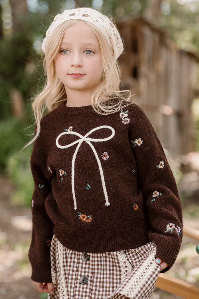 A young girl with blonde hair wears a cream knit hat and the Ivy City Co Mini Ellis Sweater in Warm Cocoa Lovelorn Ditsy, featuring floral embroidery, a bow design, and charming balloon sleeves. She completes her look with a brown gingham skirt as she stands outdoors in front of a rustic wooden structure.
