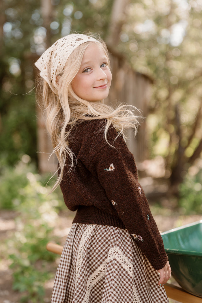 A young girl with long blonde hair, dressed in the Ivy City Co Mini Ellis Sweater in Warm Cocoa Lovelorn Ditsy—with its charming balloon sleeves and delicate embroidered details—pairs it with a checkered skirt and a crocheted headscarf. She stands outdoors, smiling over her shoulder amid lush greenery and gentle sunlight.