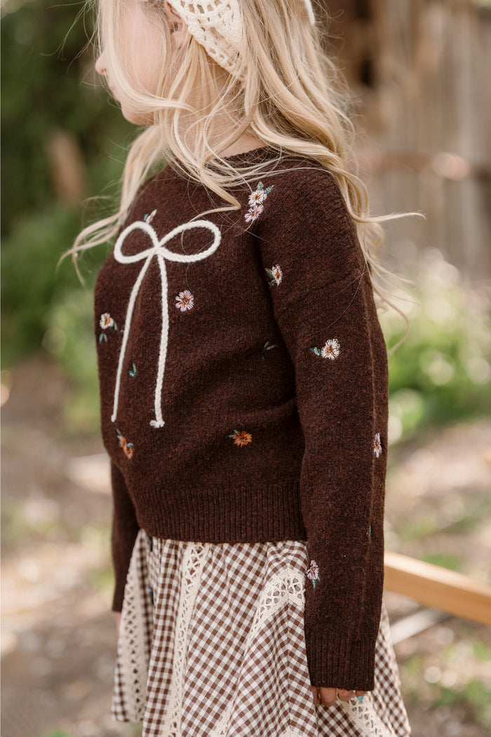 A young girl wearing the Ivy City Co Mini Ellis Sweater in Warm Cocoa Lovelorn Ditsy—with embroidered details, balloon sleeves, white bow and floral designs—paired with a patterned skirt and headband, stands outdoors among greenery and a wooden structure.