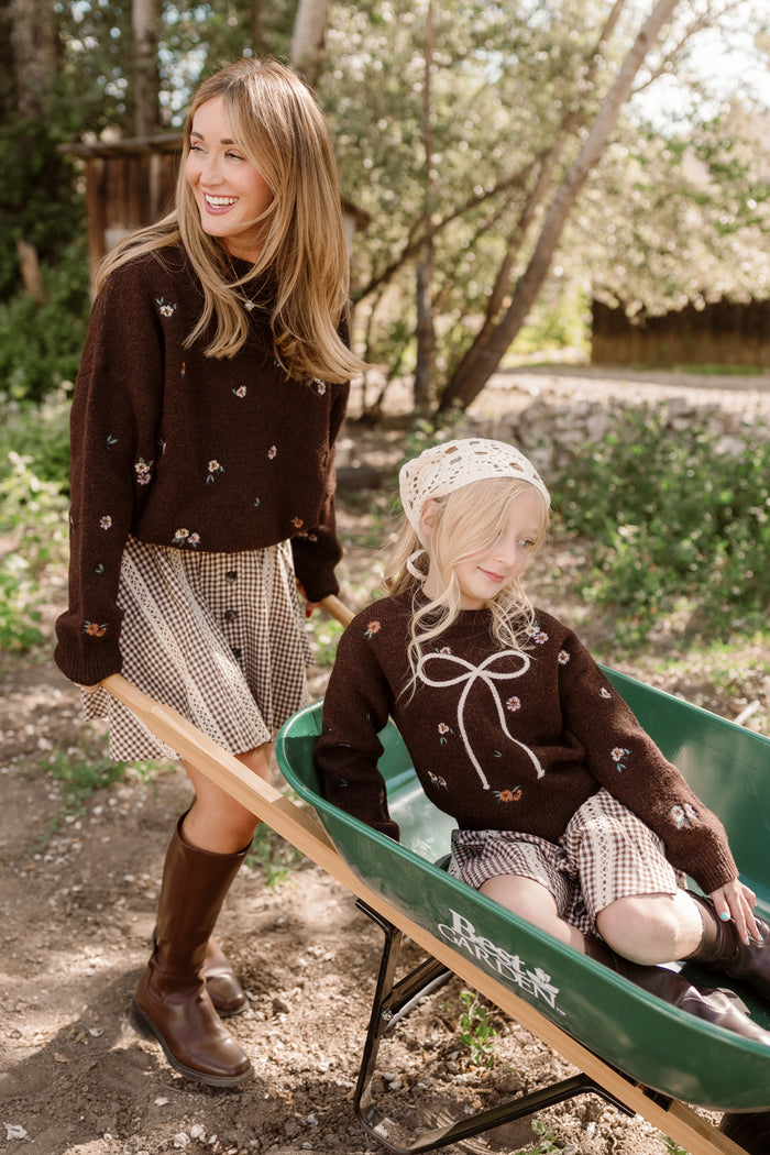 A woman and a young girl, both wearing the Ivy City Co Mini Ellis Sweater in Warm Cocoa Lovelorn Ditsy with subtle embroidered details and checkered skirts, smile outdoors. The girl's sweater features charming balloon sleeves, and she also wears a knit bonnet as she sits in a green wheelbarrow while the woman stands beside her. Trees and sunlight fill the background.